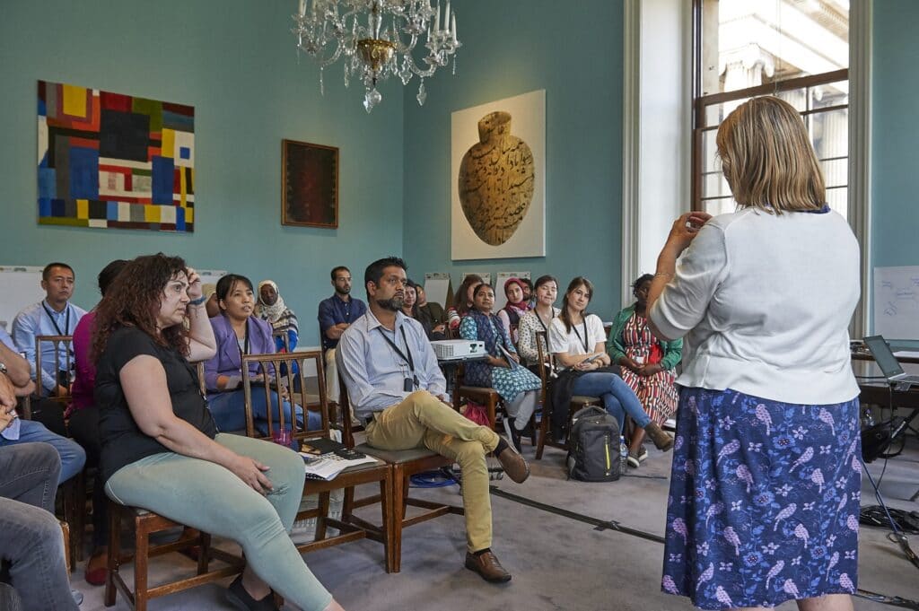 A woman stands facing a room full of people sat down in chairs. The women standing is speaking to group as they listen.