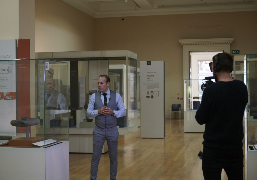 Lotfi stands next to a display case during the filming of his talk. The cameraman holds a camera facing Lotfi.