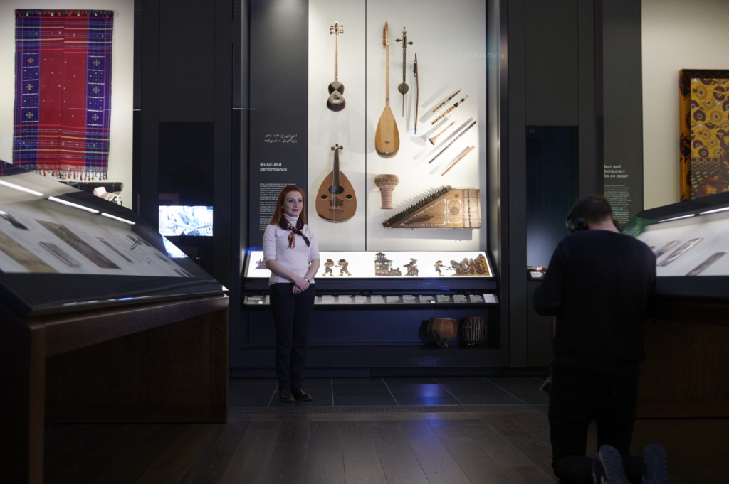 Tatevik Saroyan stands in front of a display case preparing to film her presentation. The cameraman faces away from the photo holding a lights towards Dana.
