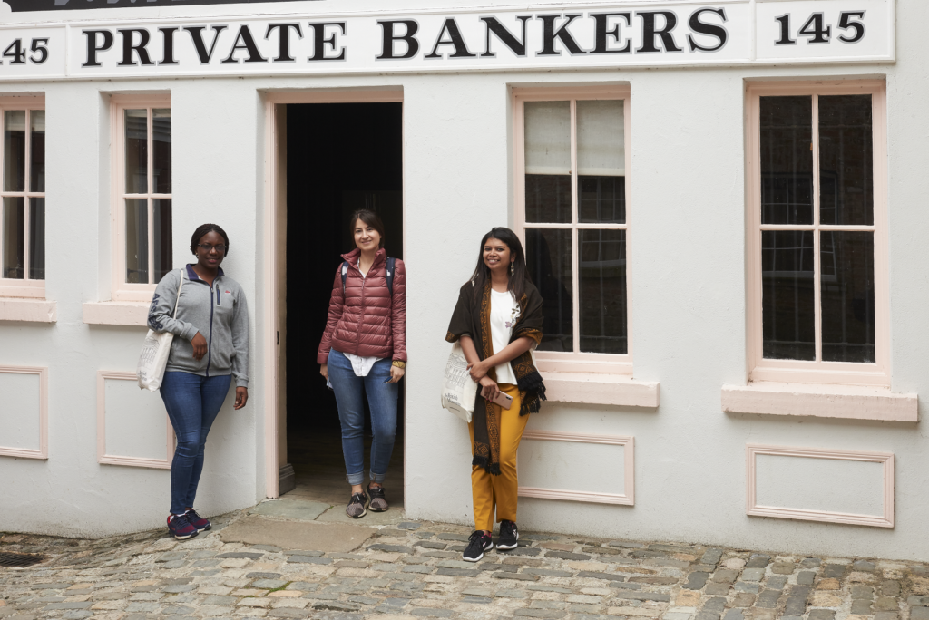 Namrata with two colleagues stand under a sign which reads 'Private Bankers' at the Ulster American Folk Park, Northern Ireland