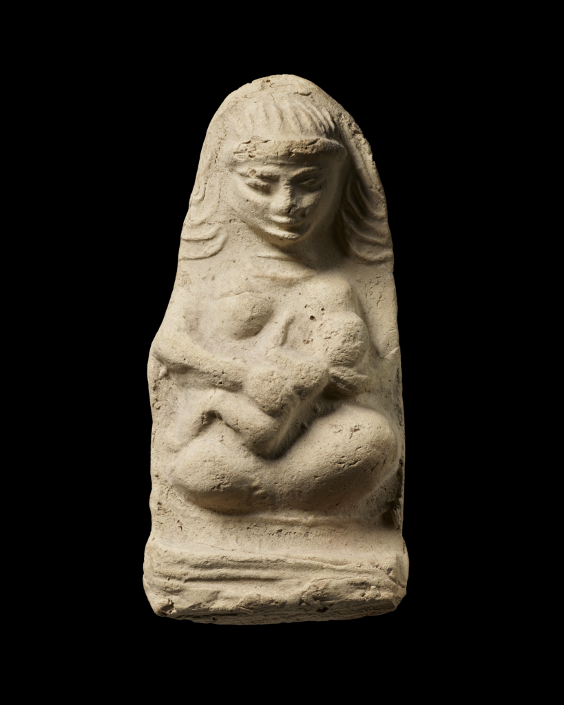 Baked clay figurine of woman and child in relief. Woman is seated cross-legged with the child on her lap suckling at her left breast