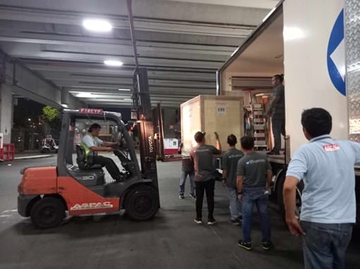 Photograph of a man driving a forklift truck, lifting a crate from a container whilst people watch.