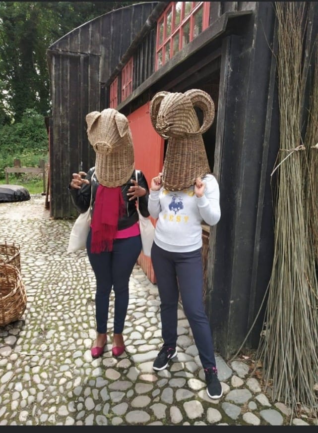Two women stand side by side holding wicker animal heads over their heads and shoulders.
