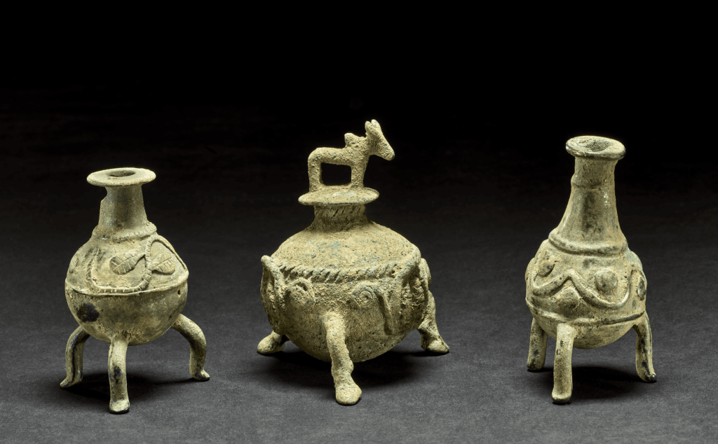 Copper-alloy cosmetic container; cast; spherical pot on three bull's legs, three leaves held up by scrolling between legs; coiled cable around vessel body; neck and mouth at top of vessel, stopper with lid in the form of a standing humped bovine.
